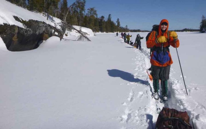 a line of cross country skiers, each pulling small sleds, make their way across a frozen, snowy lake